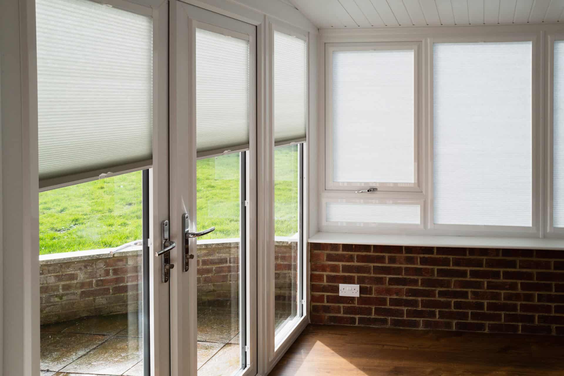 Made to measure blinds on windows and doors in a white conservatory with wooden floor and red brick walls on a sunny day. The roof is an insulated warm roof with mock beams.